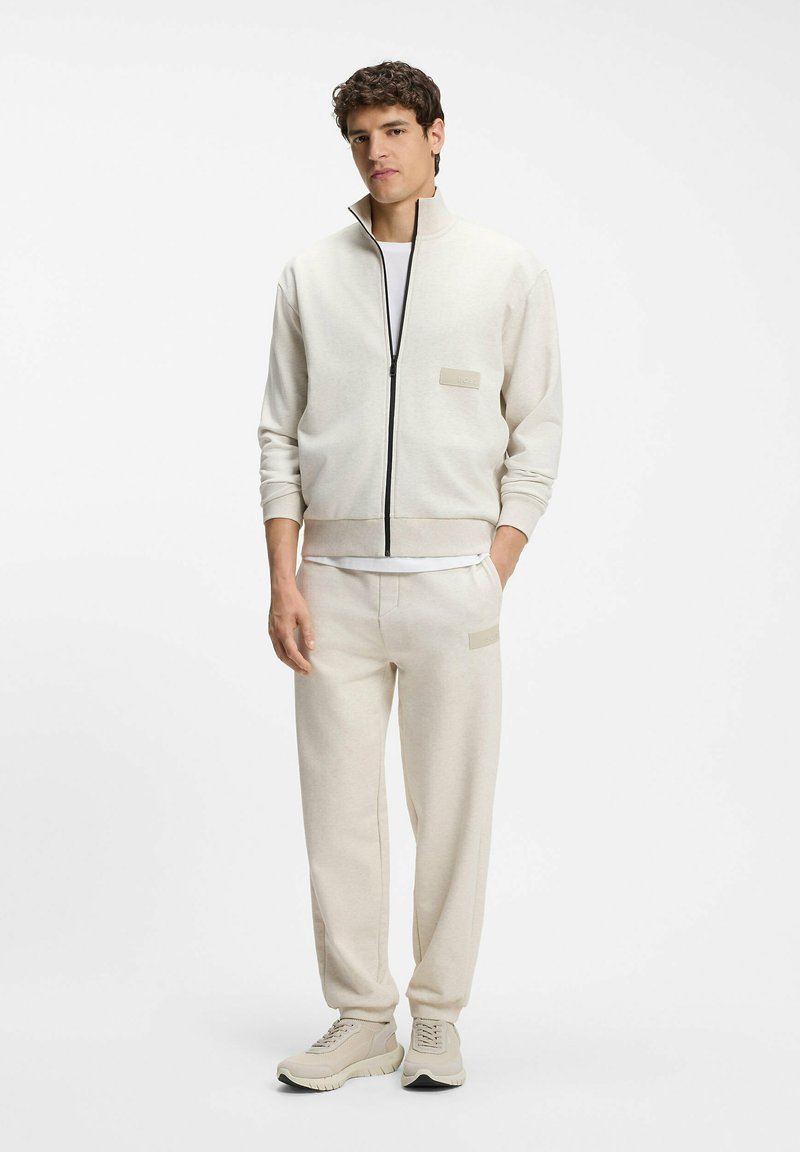 Jeune homme debout les mains dans les poches, portant une veste à fermeture éclair beige, un pantalon assorti, un t-shirt blanc et des baskets beiges sur fond blanc.