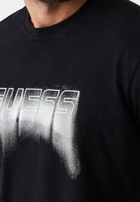 Černé bavlněné tričko s velkým, bílým graffiti nápisem "GUESS" na hrudi, který má efekt vyblednutí a vzor malých teček.