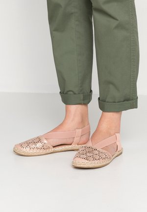 Stopy w jasnoróżowych kwiecistych balerinach slingback z wycięciami, połączone z podwiniętymi oliwkowo-zielonymi spodniami na jasnej podłodze.
