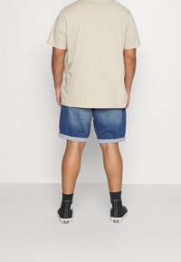 Beige puuvillane T-särk, sinised teksashortsid keeratud varrukatega, mustad kõrge säärega spordijalatsid ja mustad sokid, vaatega seljalt.
