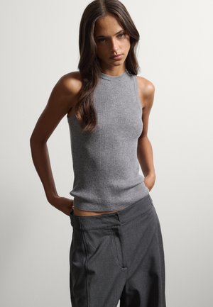 Giovane donna con lunghi capelli castani che indossa un top grigio senza maniche a coste e pantaloni grigio scuro a vita alta, in piedi davanti a uno sfondo semplice.