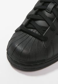 Detail černé sportovní boty, vyrobené z texturovaného syntetického materiálu, s oblým špičatým tvarem a vroubkovaným vzorem, a černými tkaničkami na hladkém povrchu.