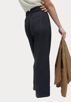 Pantaloni in denim scuro con vita alta, gambe larghe e tasche posteriori, caratterizzati da dettagli di cuciture gialle. Una giacca marrone è tenuta in una mano.