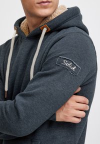 Hoodie gris foncé avec une doublure en fausse fourrure beige, doté d'un cordon de serrage, d'accents en cuir et d'un patch avec le texte "Solid" sur la manche.