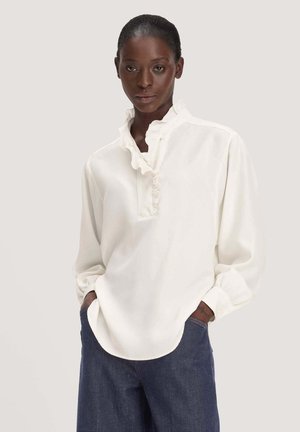 Vrouw draagt een witte blouse met lange mouwen en een geplooide kraag en donkerblauwe spijkerbroek, staand met handen in de zakken tegen een effen achtergrond.