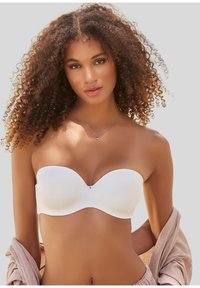 Sieviete ar cirtainiem matiem, valkājot baltu strapless krūšturi un gaiši bēšu kreklu, kas nokārts no pleciem, ar vienkāršu fonu.