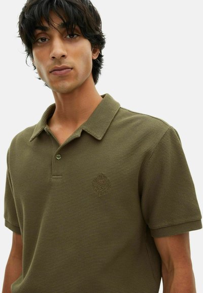 Giovane uomo con capelli scuri che indossa una polo corta di colore verde oliva con una texture e un logo ricamato sottile sul petto.