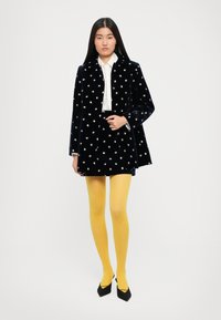 Kobieta ubrana w czarny aksamitny płaszcz i spódnicę z małymi wzorami kwiatowymi, białą bluzkę, jaskrawe żółte rajstopy i czarne spiczaste szpilki, stoi.