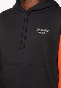 Sudadera negra con acentos naranjas, que cuenta con un bolsillo canguro y cordones ajustables. Logotipo bordado en blanco de "Calvin Klein Jeans" en el pecho.