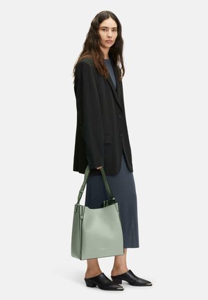 Miętowozielona skórzana torba w kształcie wiadra z regulowanymi paskami, gładką fakturą i minimalistycznym designem. W połączeniu z czarnym żakietem i ciemną sukienką.
