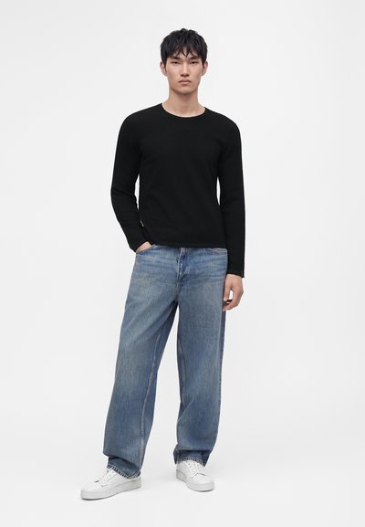 Jeune homme debout portant une chemise noire à manches longues, un jean bleu ample, des baskets blanches, une main dans la poche, sur un fond uni.