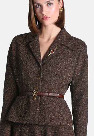 Femme portant un blazer brun texturé boutonné avec une ceinture ajustée à la taille et une broche dorée, associé à des boucles d'oreilles torsadées dorées.