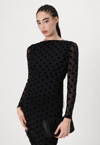 Μαύρο φόρεμα με μακριά, διάφανα μανίκια, που διαθέτει σχέδιο με πουά και συγκεντρωμένη σχεδίαση. Συνδυάζεται με μια μαύρη, υφασμάτινη τσάντα.