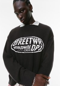 Mørkebrune strikkede trøje med hvid grafisk tekst "STREETWR WORLDWIDE CREW" på forsiden; løs pasform, ribbede manchetter, lagdelt over en ternet skjorte.