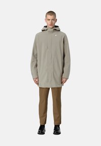 Cappotto impermeabile grigio chiaro con colletto alto e cappuccio fissato. Presenta una chiusura lampo frontale, tasche laterali e un piccolo logo sul petto.