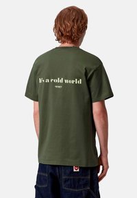 Camiseta de algodón verde aceituna con texto blanco en la parte posterior que dice "Es un mundo frío" y una etiqueta de marca visible en el dobladillo.