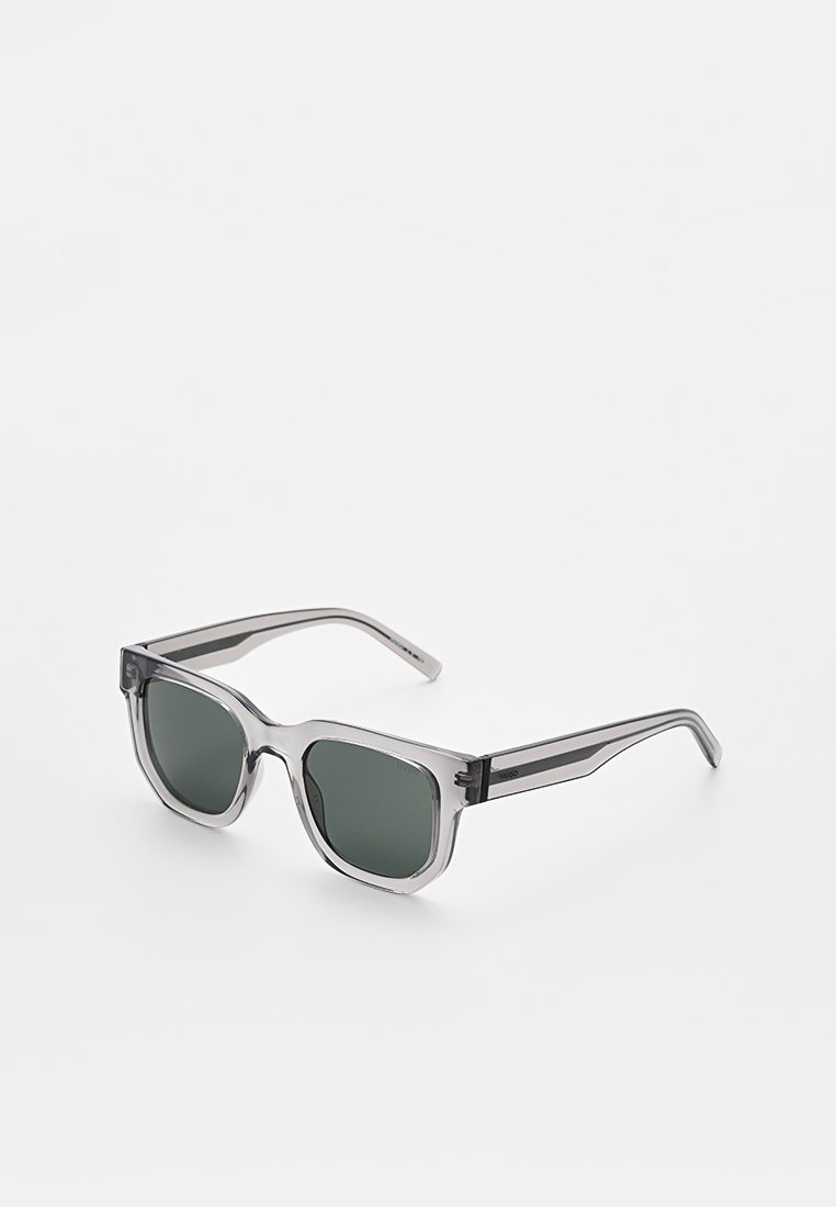 Graue transparente Sonnenbrille mit dunkelgrünen Gläsern, quadratischer Form, markantem Rahmen-Design und Metallelementen an den Scharnieren.