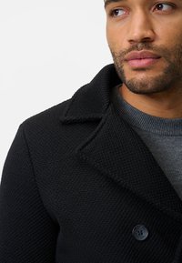 Schwarzer strukturierter Mantel mit Reverskragen und einem grauen Pullover darunter. Verfügt über einen einzelnen Knopf am Kragen und feine Nähdetails.