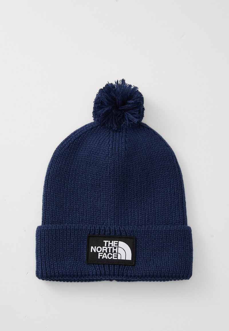 Marineblå strikket lue med pom-pom og brettet kant, utstyrt med «The North Face» logo merket i svart og hvitt.