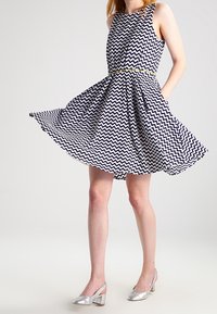 Robe à motifs zigzag noir et blanc avec une jupe évasée, un décolleté rond et une ceinture dorée. Escarpins argentés.