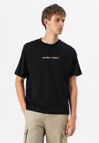 Schwarzes Baumwoll-T-Shirt mit kurzen Ärmeln, auf der Vorderseite mit einem goldbedruckten Logo "atelier dept." in der Mitte. Standardpassform.