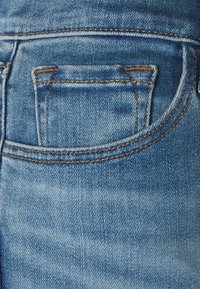 Tejido de mezclilla en azul claro con un bolsillo trasero visible y costuras marrones en contraste, mostrando una textura ligeramente desgastada y un acabado suave.