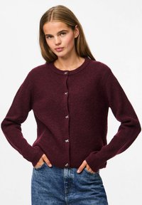 Burgundijas krāsas ribota adīta(!) kardigāns ar gariem piedurknēm, apaļu kakla izgriezumu un sudraba pogu akcentiem. Valkāts kopā ar ziliem džinsiem.