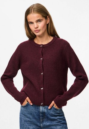 Burgunderfärgad ribbad stickad cardigan med långa ärmar, rund hals och silverknappdetaljer. Bärs tillsammans med blå denimjeans.