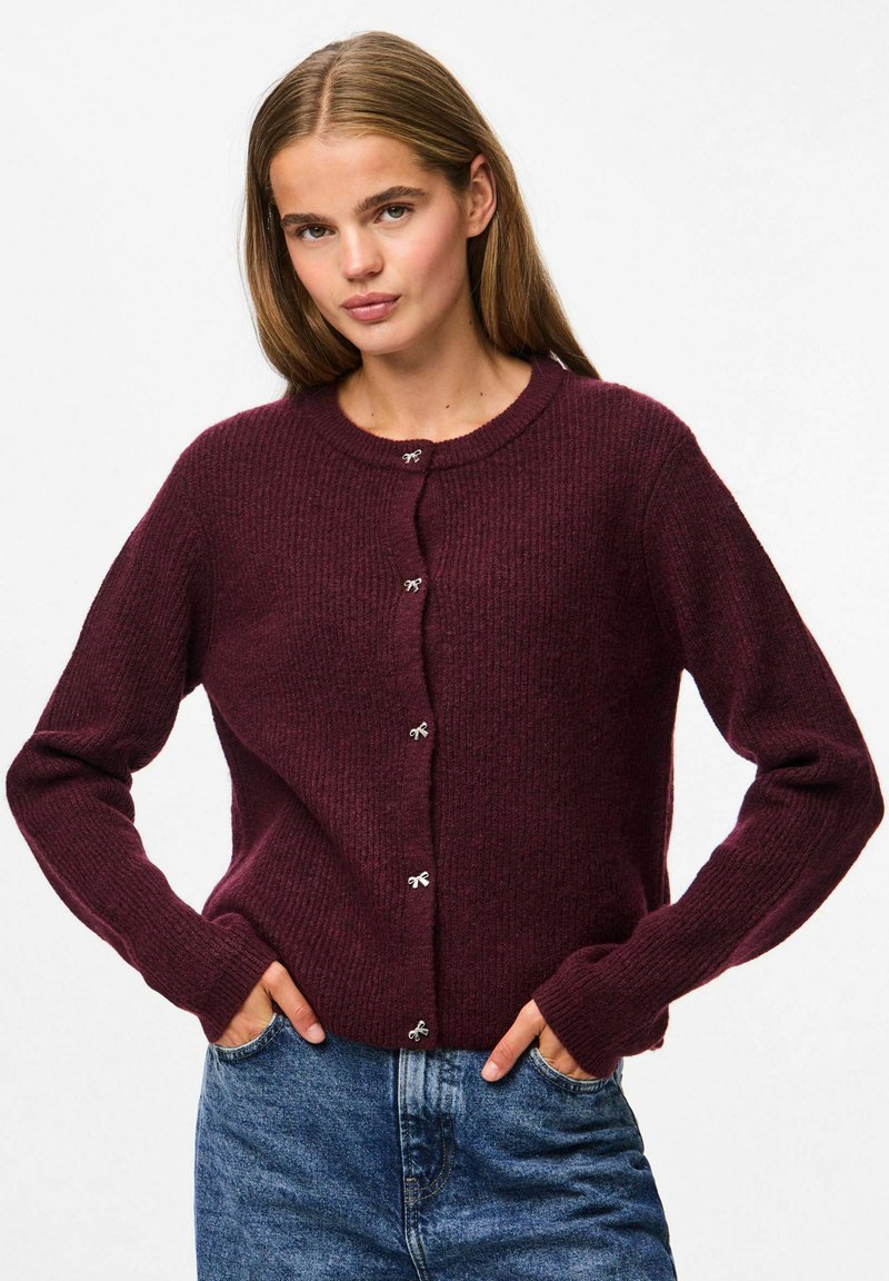 Burgundijas krāsas ribota adīta(!) kardigāns ar gariem piedurknēm, apaļu kakla izgriezumu un sudraba pogu akcentiem. Valkāts kopā ar ziliem džinsiem.