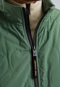 Zelena prešita jakna z visokim ovratnikom, črnim zadrgo in rumenim logotipom. Pod njo je vidna krema barva rebrasta turtleneck.