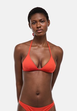 Femme aux cheveux courts portant un haut de bikini orange à col halter et un bas assorti, debout devant un fond gris clair uni.