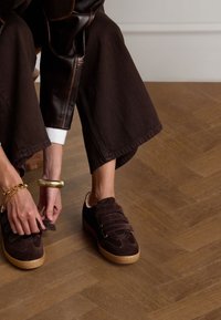 Braune Wildleder-Sneaker mit drei Klettverschlüssen und einer hellen Gummisohle. Getragen mit dunklen, weit geschnittenen Hosen auf einem Holzboden.