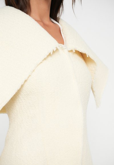 Vestido texturizado crema com um colar largo e detalhado. O tecido possui um padrão tecido subtil e bordas brutas, destacando o seu design único.