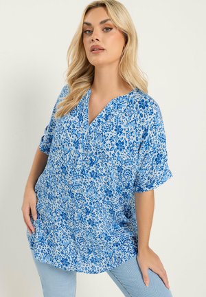 Femme blonde portant un chemisier bleu à fleurs ample à manches courtes et un jean bleu clair, posant avec les mains sur les hanches devant un fond uni.