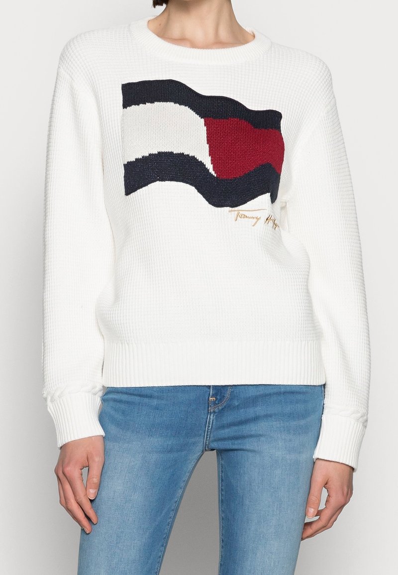 Personne portant un pull en maille blanche avec un grand motif rectangulaire rouge, blanc et bleu marine et un jean bleu clair.