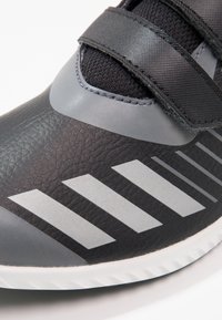 Chaussure de sport noire avec une tige texturée, des accents gris, deux bandes réglables et une semelle en caoutchouc blanche ornée de rayures grises angulaires.