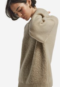 Beżowy sweter z prążkowanymi mankietami i teksturowanym, puszystym materiałem. Design ma luźny krój oraz prosty okrągły dekolt.