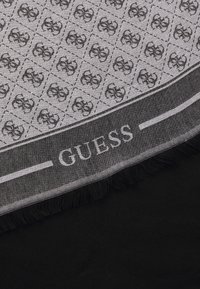 Fular alb-negru cu un model geometric și margini franjurate, etichetat cu "GUESS" pe margine. Fabricat dintr-un material ușor.