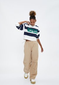 Afskåret hvid og marineblå stribet sweatshirt med grøn "California West Coast" tekst, parret med beige løse bukser og hvide sko.