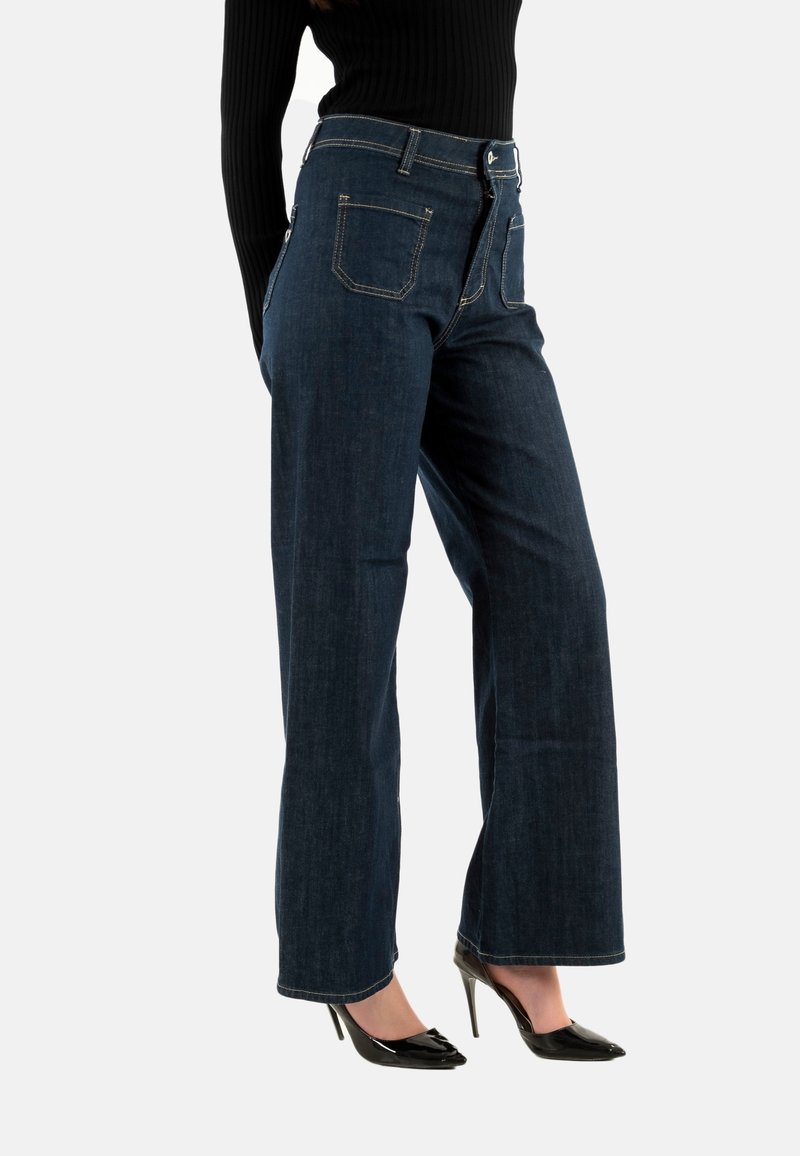 Pantalones de mezclilla anchos de color azul oscuro con cintura alta, cierre frontal con botones y bolsillos traseros. Llevados con zapatos de tacón negros.