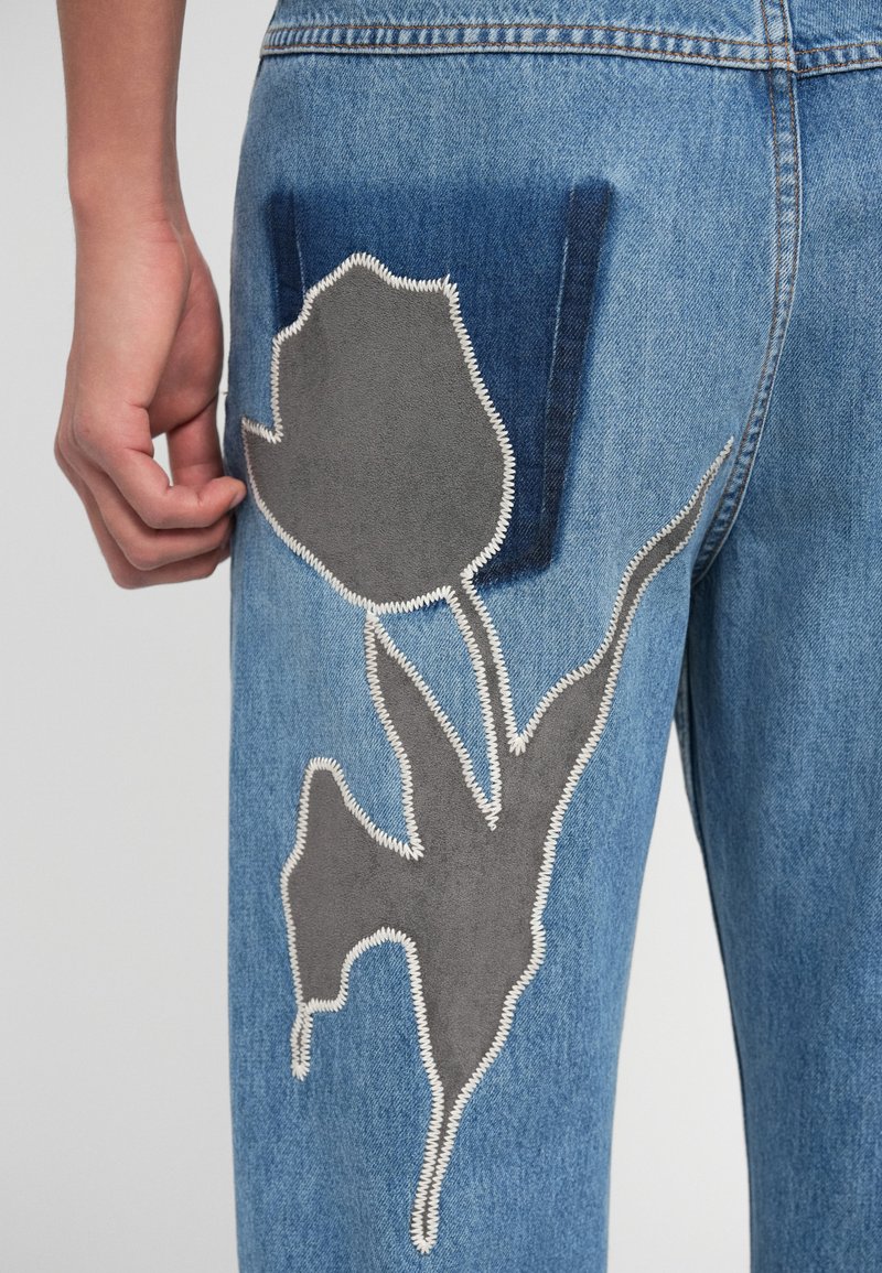Jeans aus Denim mit blauen und grauen Blumenapplikationen. Hervorhebung durch kontrastierende Nähte und ein abstraktes Design. Nahaufnahme der Gesäßtasche.
