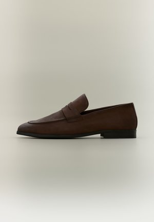 Dunkelbrauner Lederloafer mit niedrigem schwarzem Absatz und einem Riemendetail über dem Obermaterial, seitlich auf neutralem Hintergrund gezeigt.