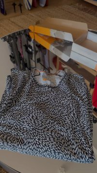 Short-sleeved shirt in black with white irregular oval patterns, featuring a rounded neckline and gathered details at the shoulders.