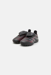Baskets de sport noires avec des accents rouges, tige texturée, sans lacets traditionnels, et semelle en caoutchouc conçue pour une traction sur les surfaces en gazon.