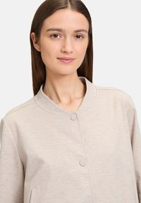 Jasnobeżowy, teksturowany kardigan z okrągłym kołnierzem, dwiema guzikami i bocznymi kieszeniami, wykonany z miękkiej mieszanki tkanin.