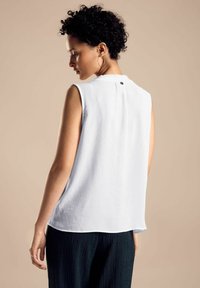 Top bianco senza maniche con finitura testurizzata, vestibilità ampia e piccolo dettaglio con bottone sul retro; abbinato a pantaloni scuri e testurizzati.