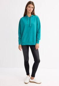 Sudadera de color turquesa de algodón con un bolsillo canguro, cordones ajustables, mangas largas y un corte holgado, combinada con jeans oscuros.