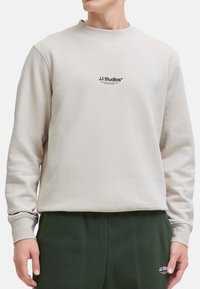 Hellbeige Sweatshirt mit Rippstruktur, Rundhalsausschnitt und schwarzem "JJ Studios" Logo. Kombiniert mit dunkelgrünen Sweatpants mit passendem Logo.