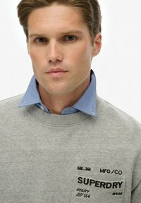 Joven con cabello corto castaño que lleva una sudadera gris de Superdry sobre una camisa de cuello azul, contra un fondo blanco.