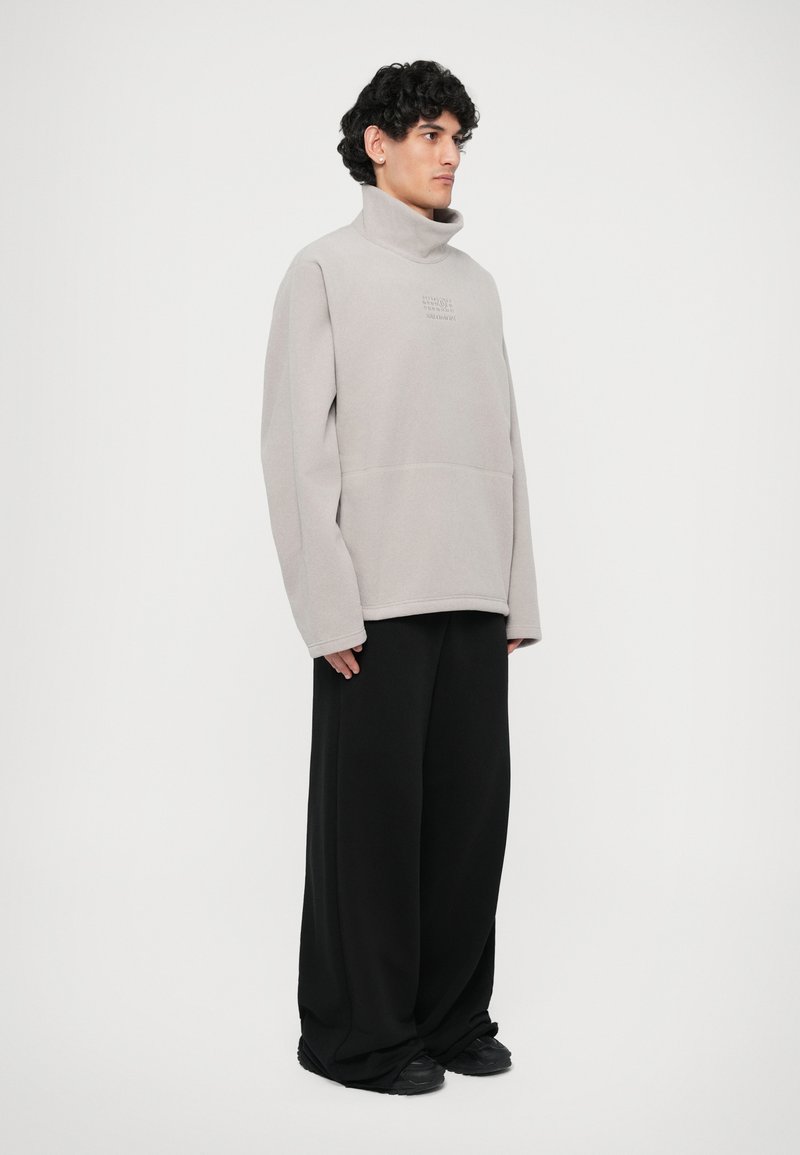 Light gray fleece pullover with a high collar and a front pocket, paired with loose black pants and black sneakers.